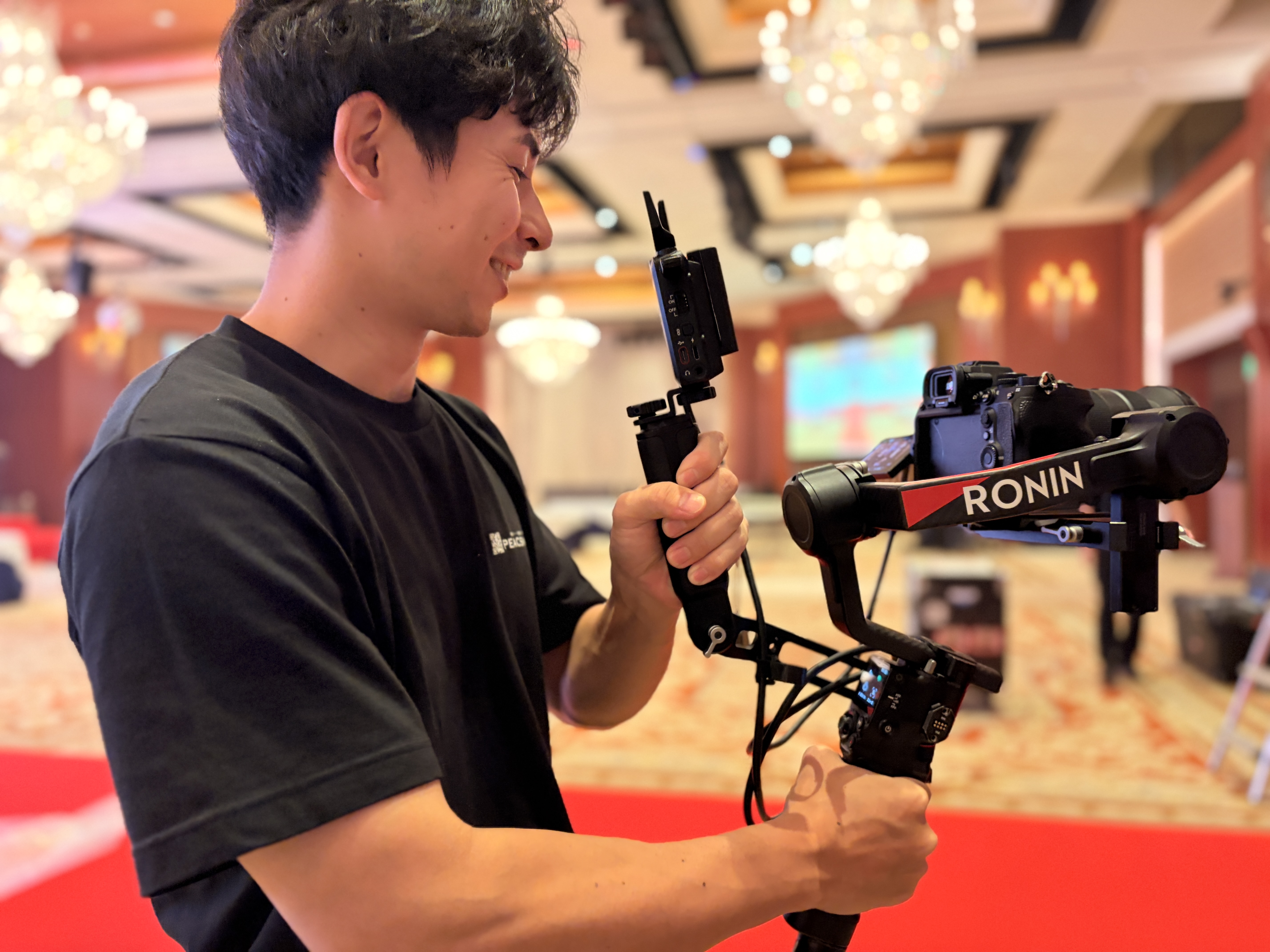 黒Tシャツの男性スタッフが、宴会場でDJI RONINジンバルを両手で構えている撮影準備の場面