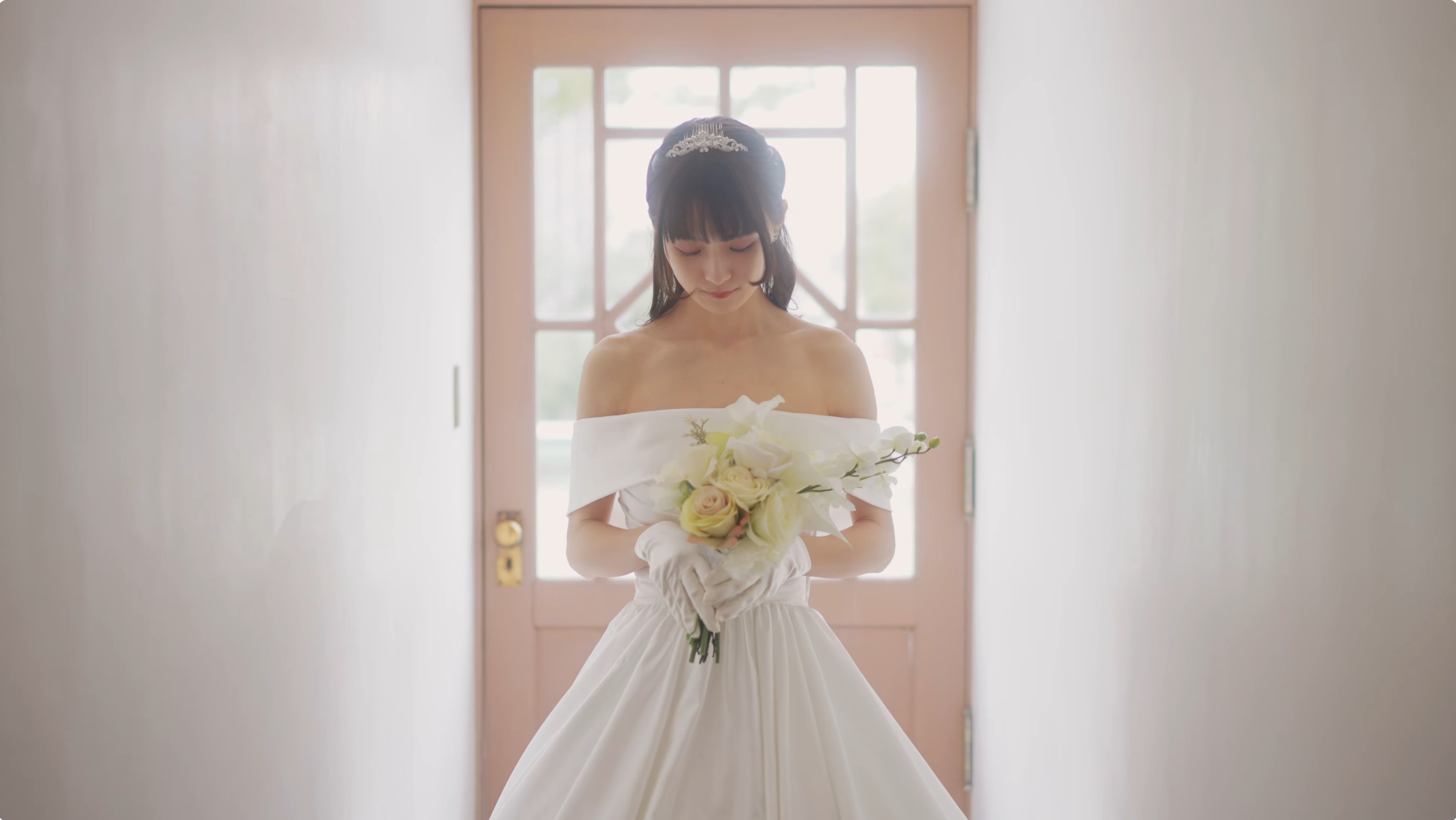 白いウェディングドレスとティアラを身に着けた女性が、白いブーケを両手で持ち、うつむいて立っている。背後にピンクのドアが見える。