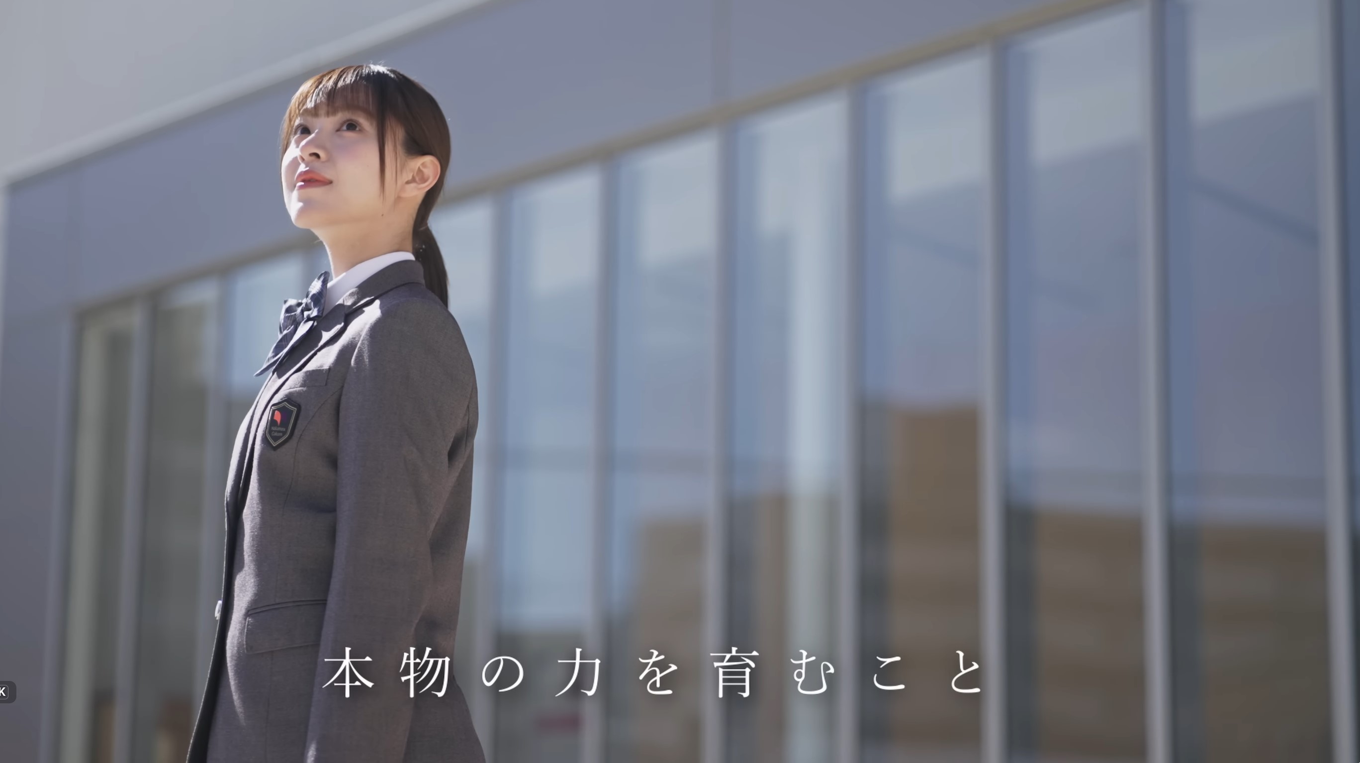 グレーの制服を着た女子生徒が、ガラス張りの建物前で上を見上げている。テロップに「本物の力を育むこと」と表示。