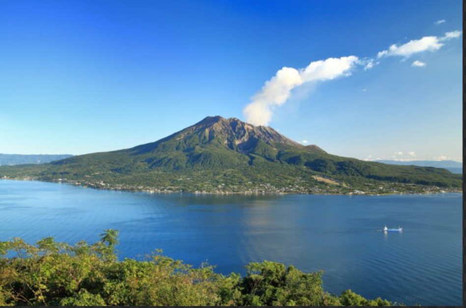 山頂から白煙を上げる火山と手前に広がる青い湾、小型船が浮かぶ晴天の風景