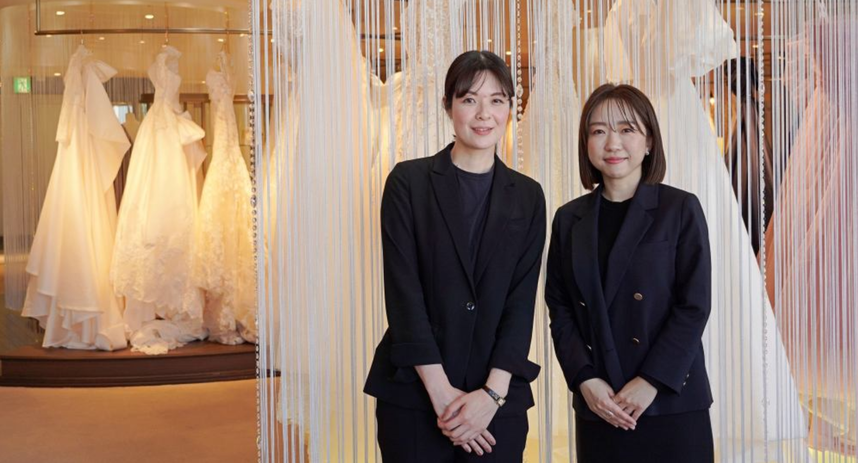 黒いスーツ姿の女性2名がウェディングドレス展示のある店内で並んで立っている
