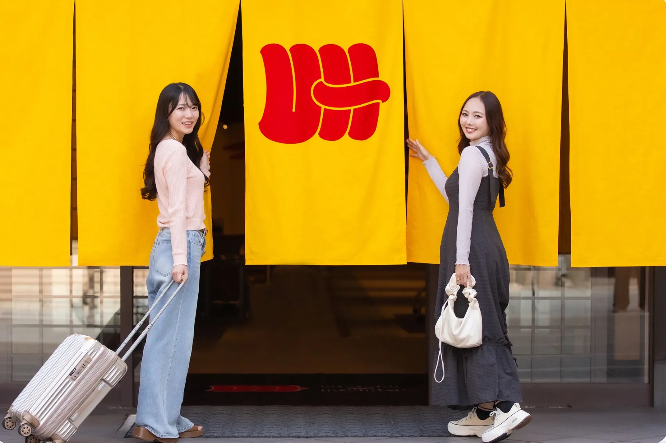 黄色い暖簾の前でカメラに微笑む2人の若い女性。一方はキャリーケースを持つ。