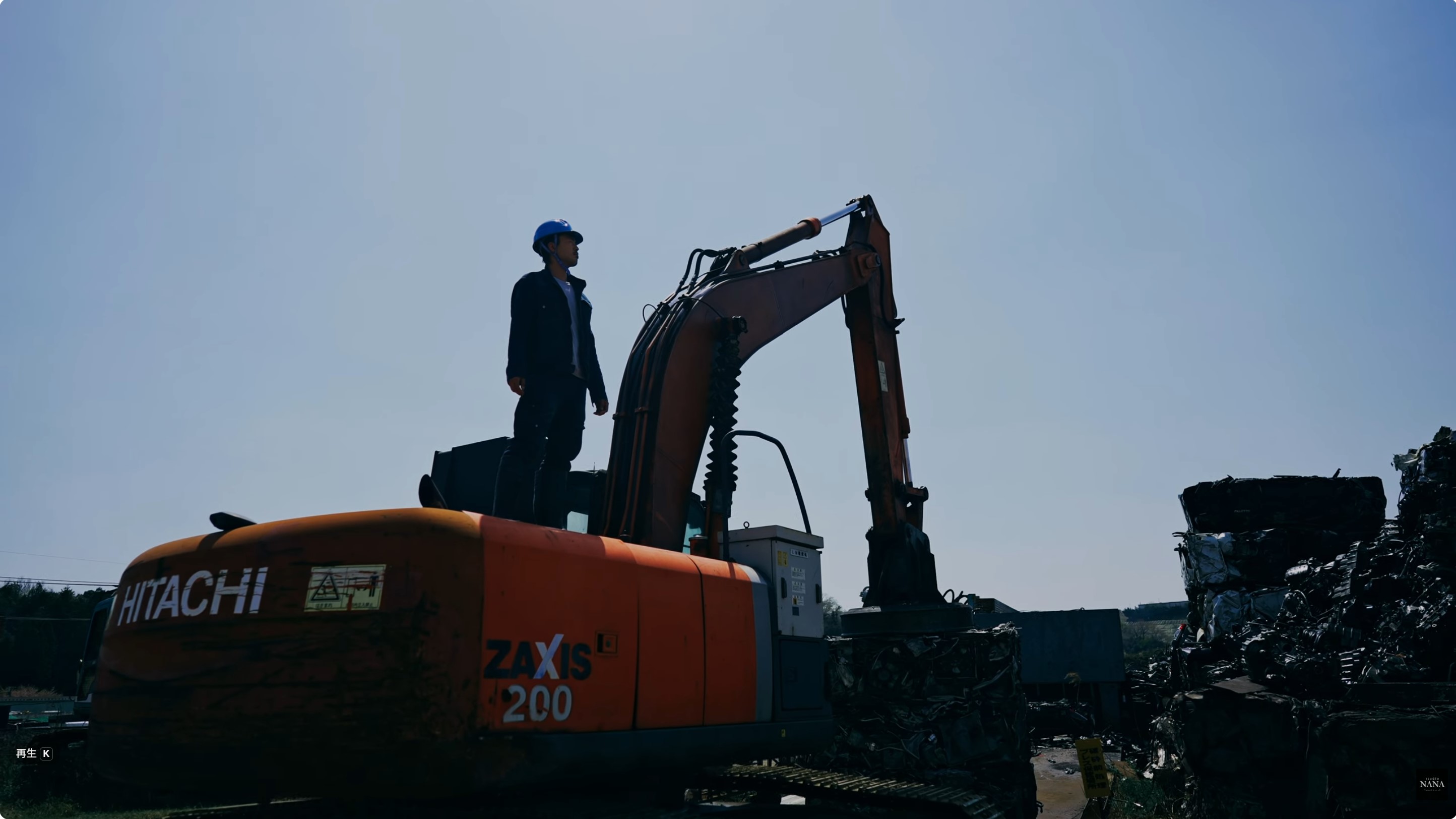 青空を背景に、大型油圧ショベルの上に立ち遠くを見渡す青いヘルメットの作業員