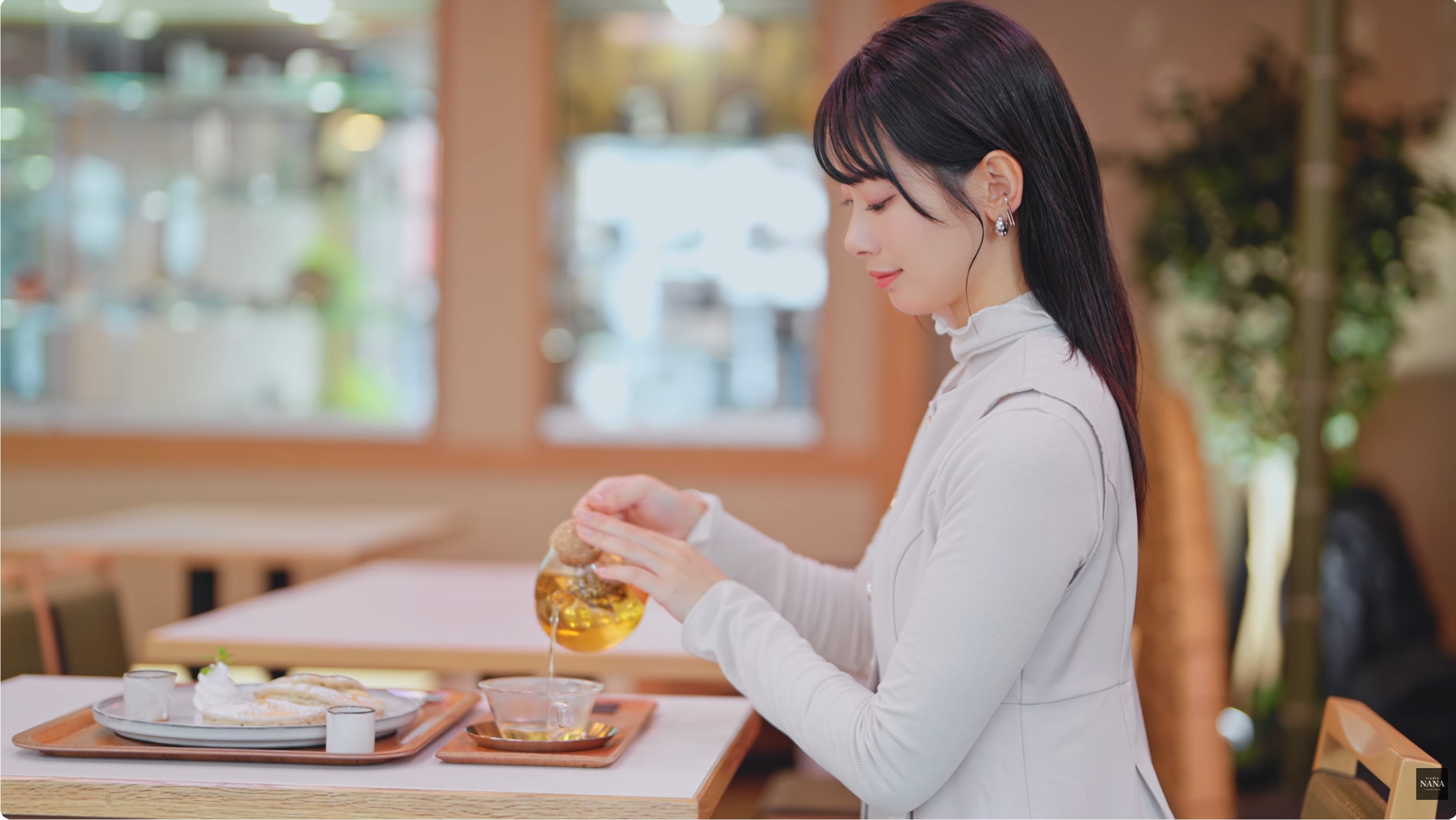 白いトップスの女性がカフェでガラスポットから茶を注いでいる場面
